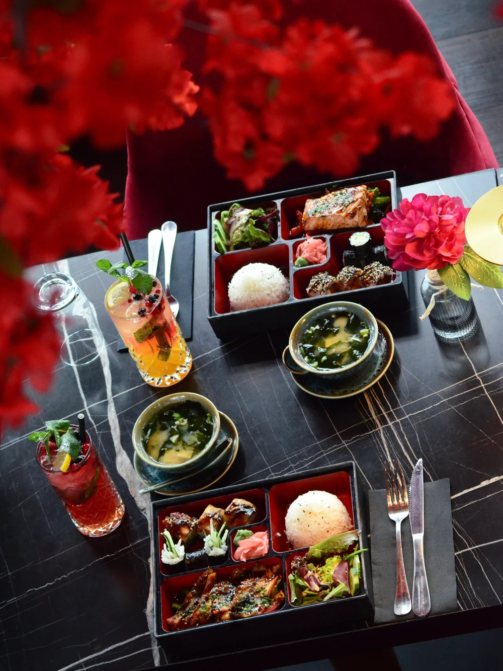 Bento-Box mit Suppe und Cocktails auf dem Marmortisch, daneben rote Blüten